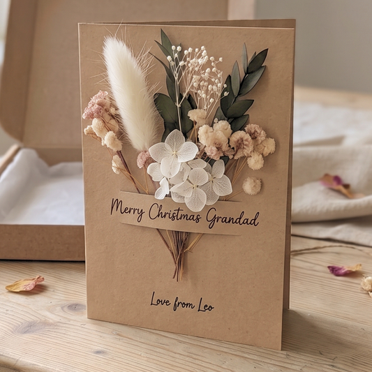 A handmade Christmas card for 'Grandad' made from brown kraft paper, decorated with dried white flowers, green leaves, and fluffy beige grasses. The card is displayed on a wooden table next to an open cardboard box.